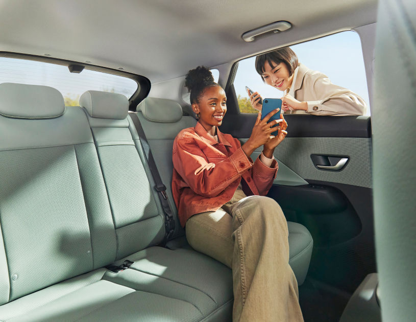 A woman sitting in the back of a The all-new KONA is looking at a smartphone screen with a woman outside the car through an open window.