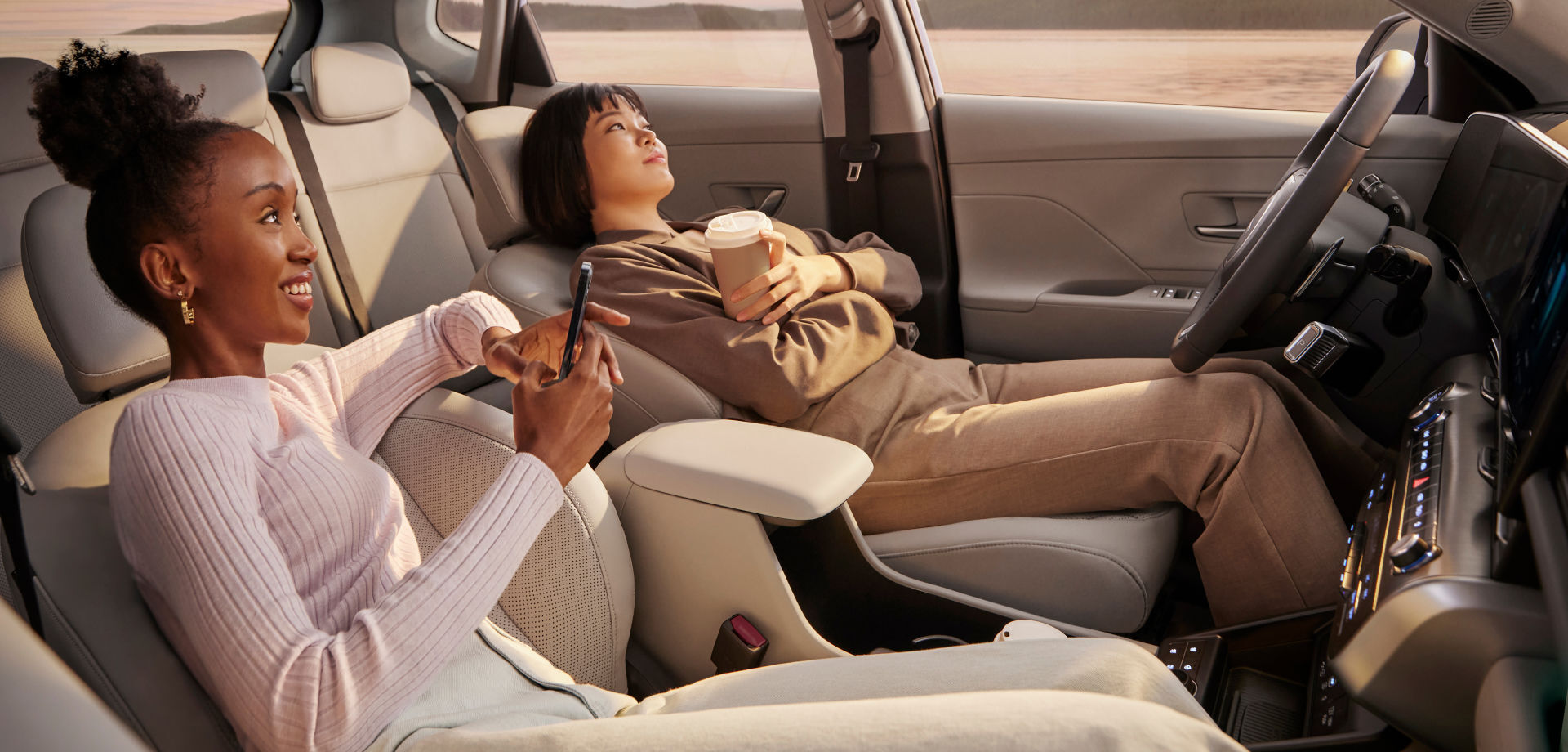 Two women are sitting in the front row of a The all-new KONA with their seats folded back, watching the sunset. The woman in the front passenger seat is holding her smartphone, and the woman in the driver's seat is holding her take-out paper cup in her left hand, with her arms crossed.