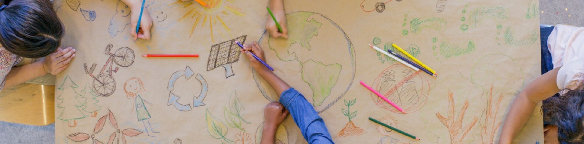Bird’s-eye view of children’s hands sketching environmentally-friendly images of Earth, solar power, etc. in colored pencils on a large paper canvas.