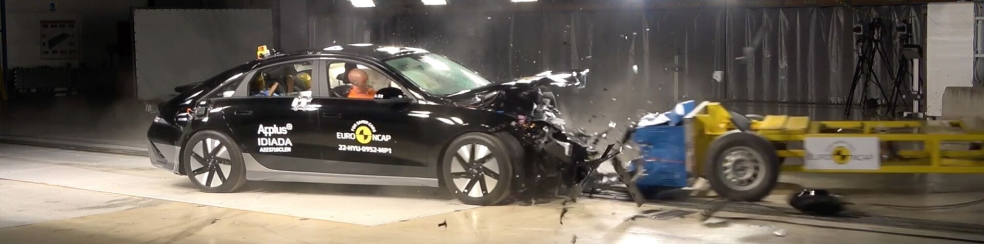 A black IONIQ 5 with 3 crash dummies inside it crashing into a yellow trailer in an empty warehouse with bright white lights as part of a safety test.