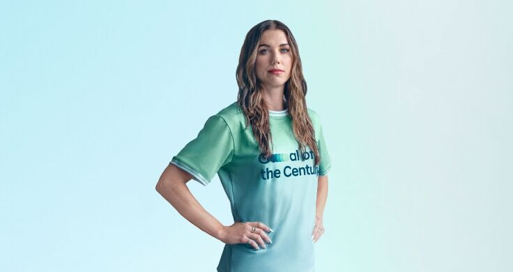 A close-up of Alex Morgan’s head and shoulders. She is wearing her green Team Century jersey and she has long wavy brunette hair. She has her hands on her hips and is looking ahead proudly.