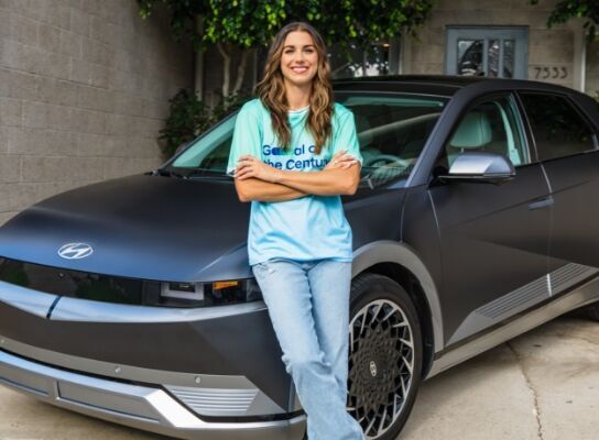 Alex Morgan standing in front of an IONIQ 5 in a garage. She is wearing her green Team Century jersey and blue jeans and has long wavy brunette hair.
