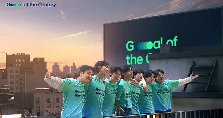 All 7 members of BTS standing on a rooftop with rooftops and a billboard with ‘Goal of the Century’ written on it behind them. All 7 members are wearing green and blue Team Century jerseys with Goal of the Century written on them.