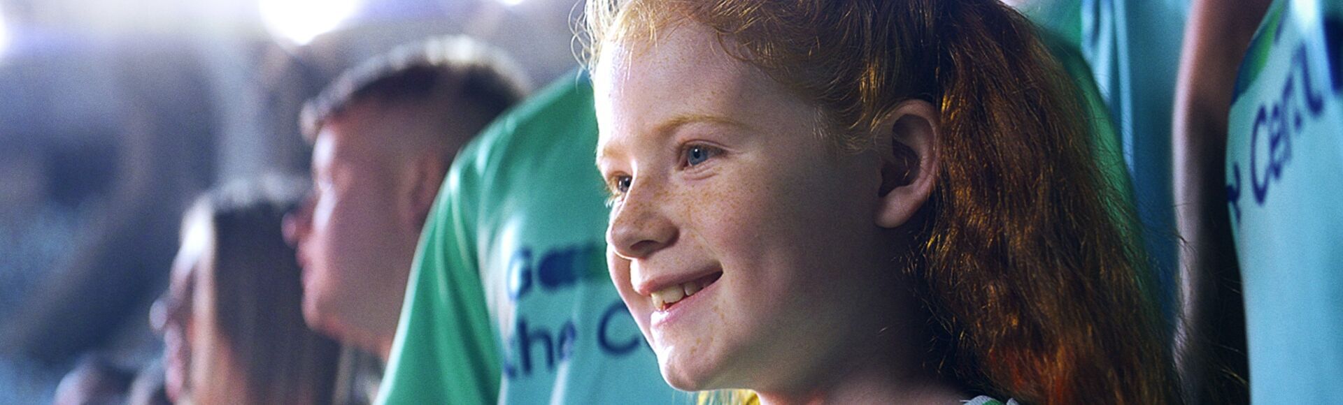 A young girl with red hair smiling surrounded by football fans with blue and green Team Century jerseys on.