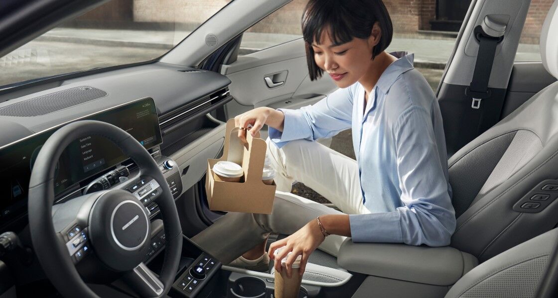 A woman is sitting in the passenger seat of The all-new KONA with the right front door open. The woman is holding a to-go drink in her right hand, and carrying her drink into a cup holder in her vehicle with her left hand.