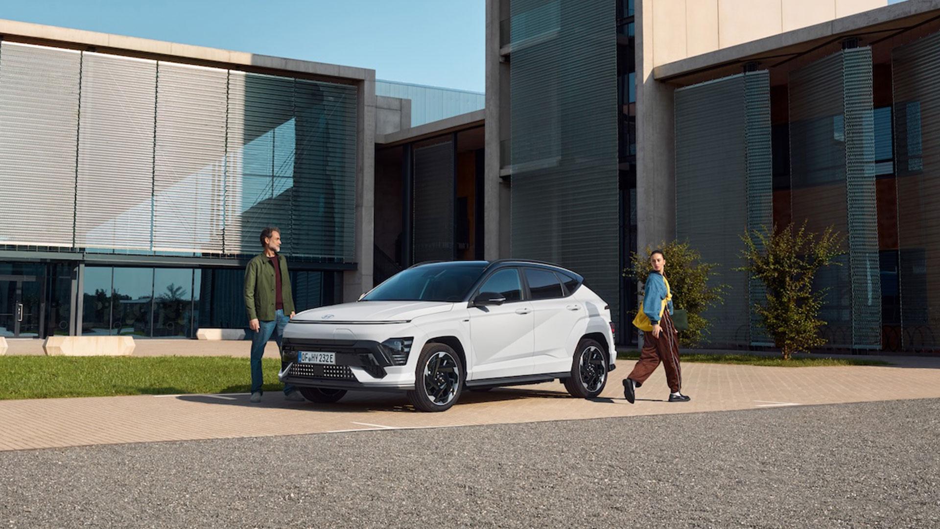 KONA Electric parkert foran en moderne bygning med to mennesker som beveger seg rundt bilen. foto.