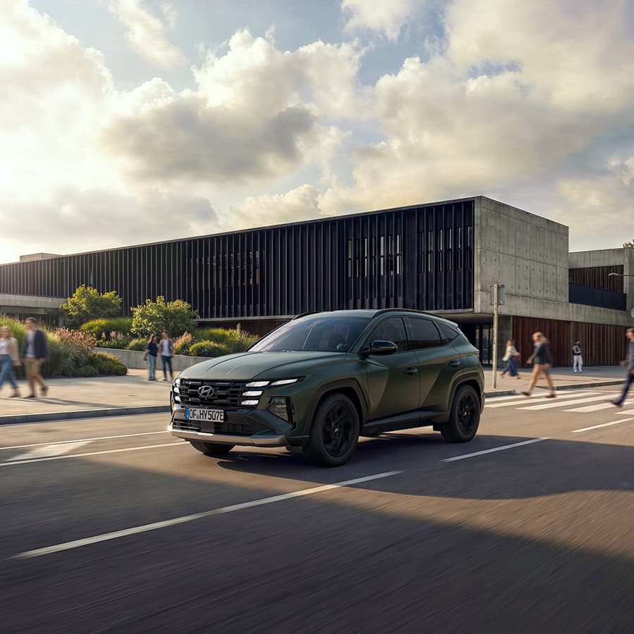 Mörkgrön Hyundai TUCSON kör förbi en modern byggnad i stadsmiljö.