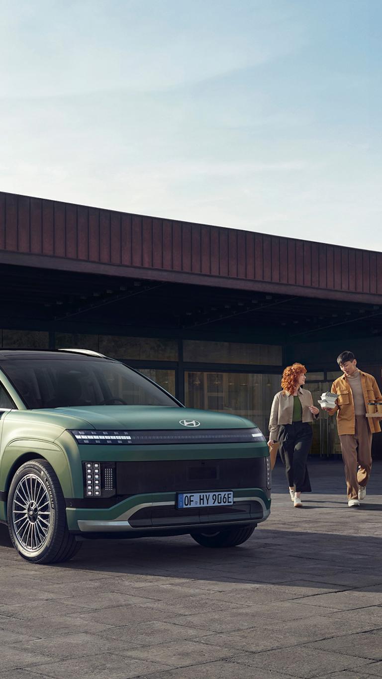 Et ungt par kommer gående med kaffe bak en IONIQ 9 som står og lader. Foto.