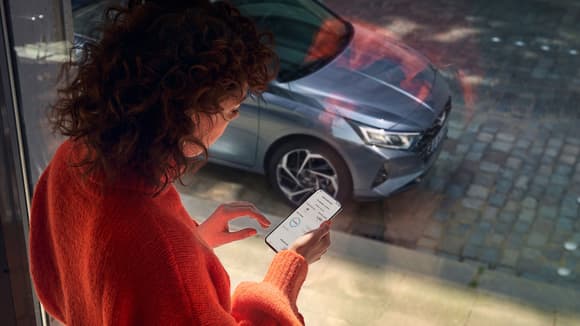 En dame står og ser på mobilen sin foran et stort vindu med en bil parkert på utsiden. Foto.