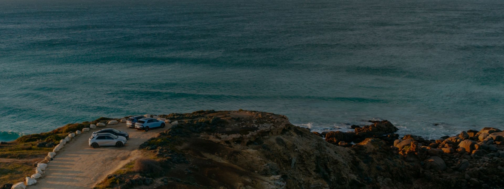 Three Hyundai cars parked on a cliffside overlooking the ocean at sunset, surrounded by rugged rocks and tranquil blue waters.