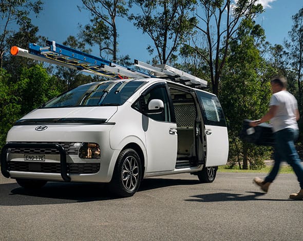 White Hyundai van fitted with roof racks and ladder parked roadside, with side door open and tools visible, highlighting fleet accessories for commercial use.