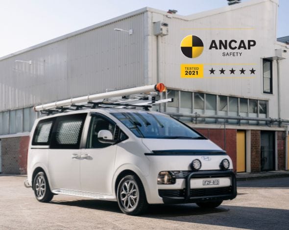Hyundai STARIA Load commercial van parked outside an industrial building, featuring roof rack accessories and a 5-star ANCAP safety rating badge, highlighting practicality, safety and fleet-ready capability.