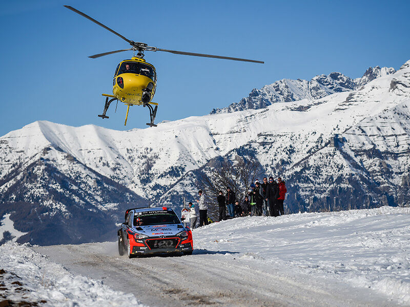 Hyundai i20 Coupe driving through snow at Monte Carlo Rally with helicopter above