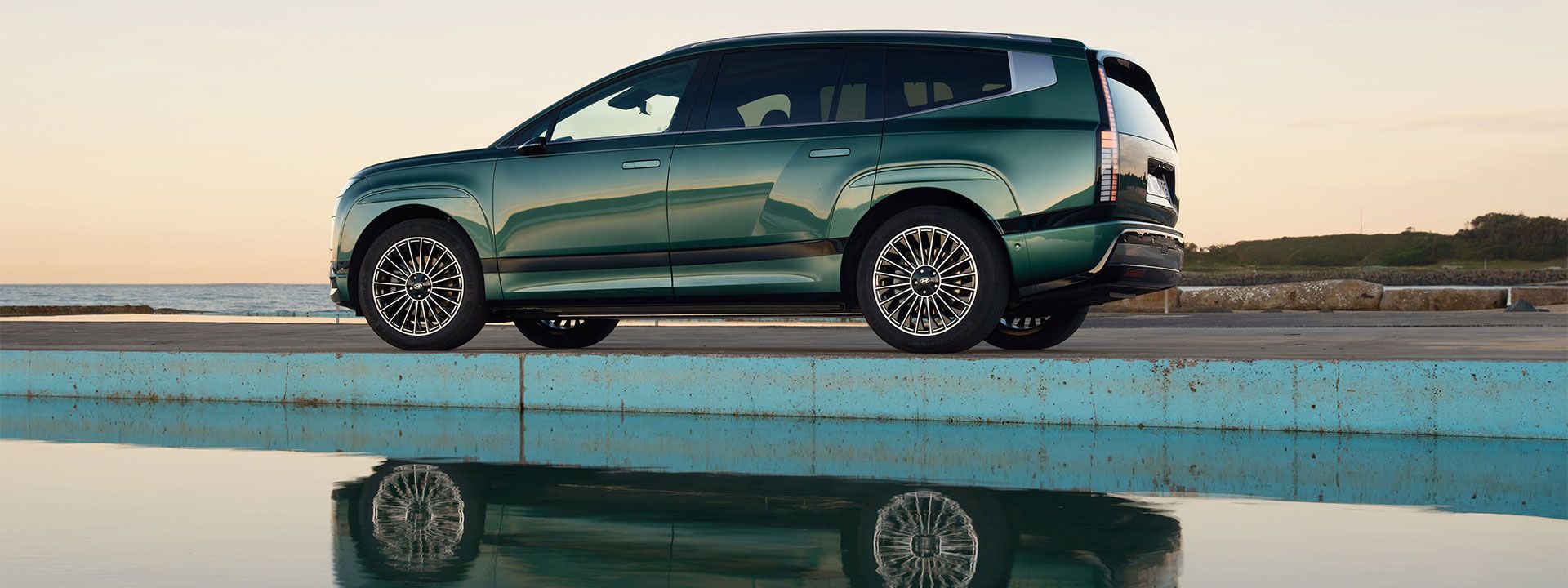 Hyundai IONIQ 9 SUV in green, reflected beside tranquil water at sunset where bold design meets serene elegance.