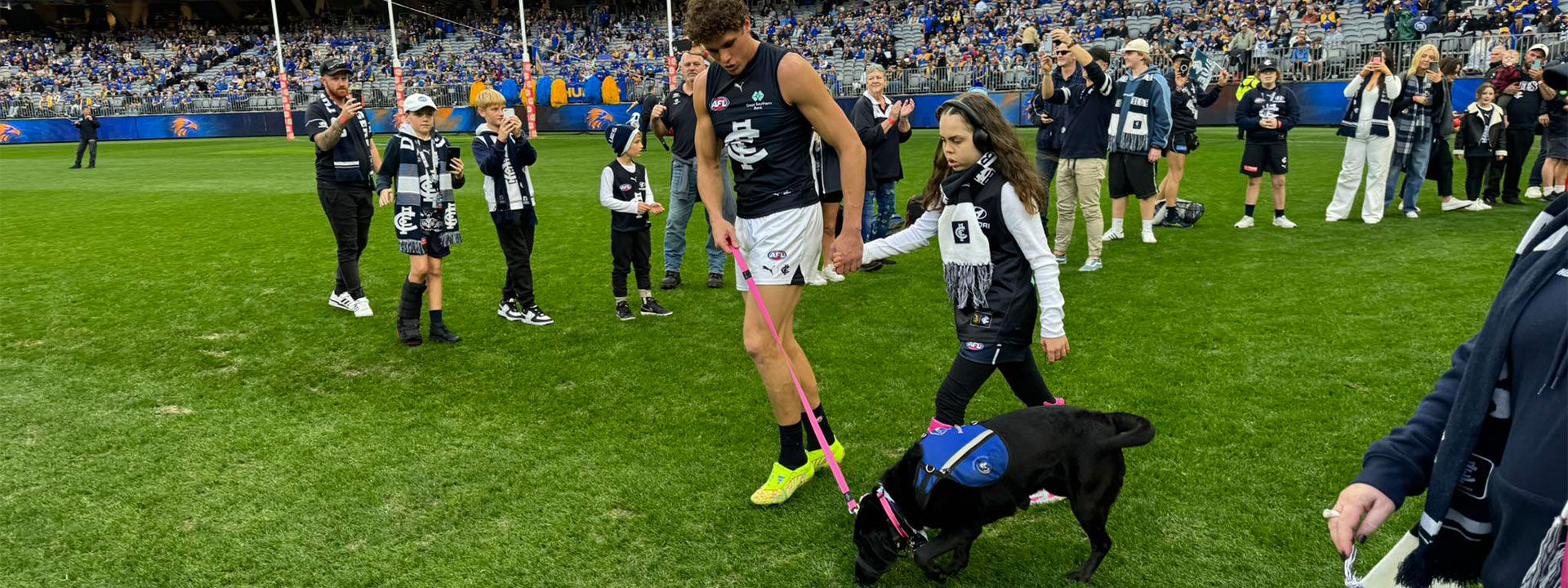 Carlton football player walking with a young girl and a black service dog on the field during a game