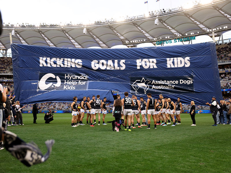 Carlton football team celebrating on field with 'Kicking Goals for Kids' banner