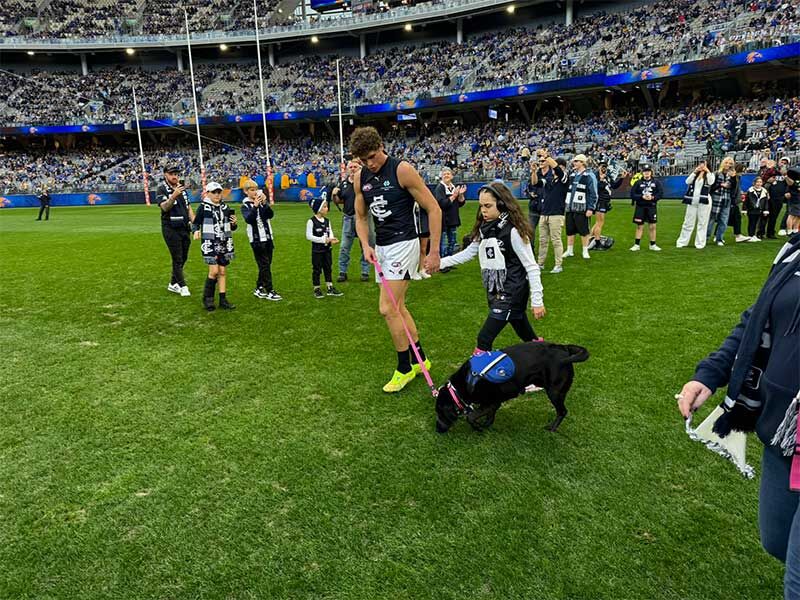 Carlton football player walking with a young girl and a black service dog on the field during a game