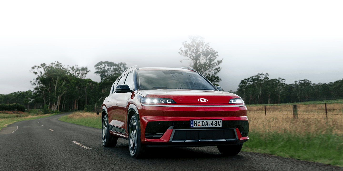 Front-facing view of a vibrant red Hyundai ELEXIO driving along a two-lane country road with its daytime running lights on, set against a backdrop of green fields, line of trees, and an overcast, misty sky.