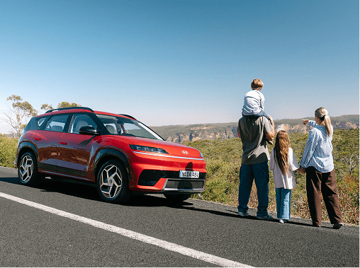 A family of four stands next to a red Hyundai ELEXIO parked on the roadside, looking out at a wide, scenic panorama of the Blue Mountains under a clear blue sky.