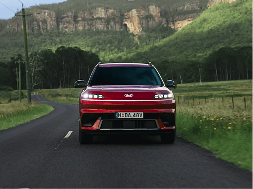 Front-facing, centered shot of a red Hyundai ELEXIO driving down a country road, highlighting its distinctive pixel-style daytime running lights against a scenic backdrop of steep, forested cliffs.