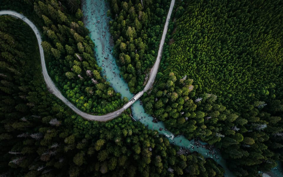 Aerial view of a winding road cutting through a dense green forest, crossing over a turquoise river via a narrow bridge, highlighting nature's untouched beauty and serene wilderness.