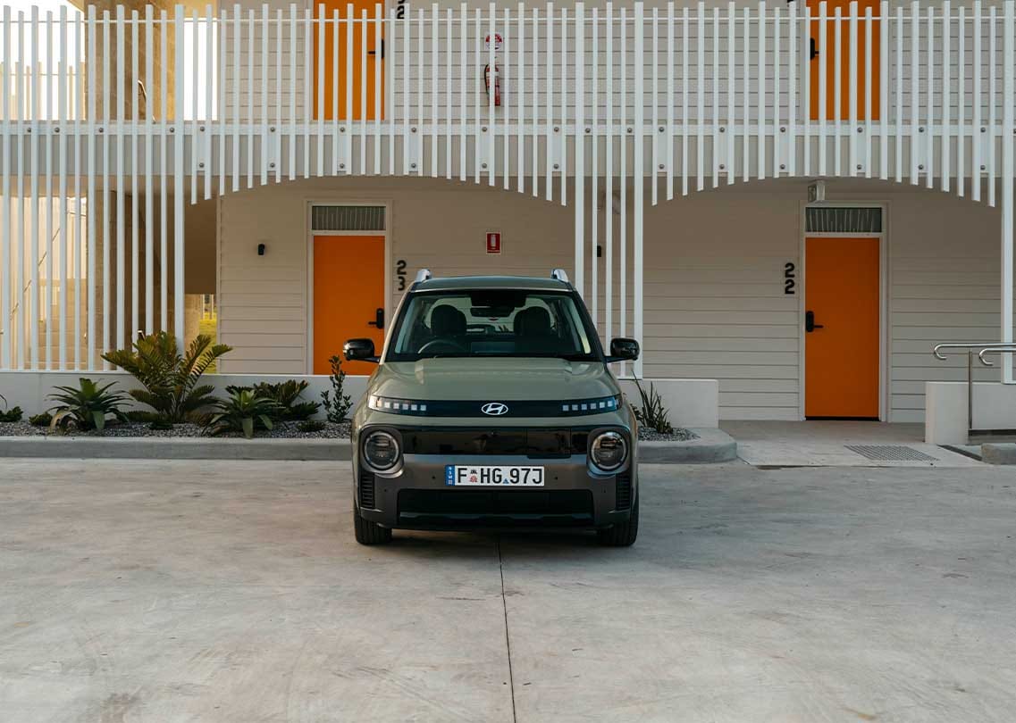 Front view of the Hyundai INSTER in Khaki, parked in a modern urban setting, showcasing its distinctive design, compact SUV proportions, and futuristic styling.