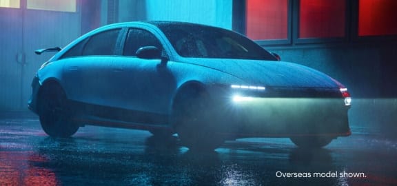 A blue Hyundai Ioniq 6 N with illuminated headlights in a dimly lit, urban setting with red and blue ambient lighting