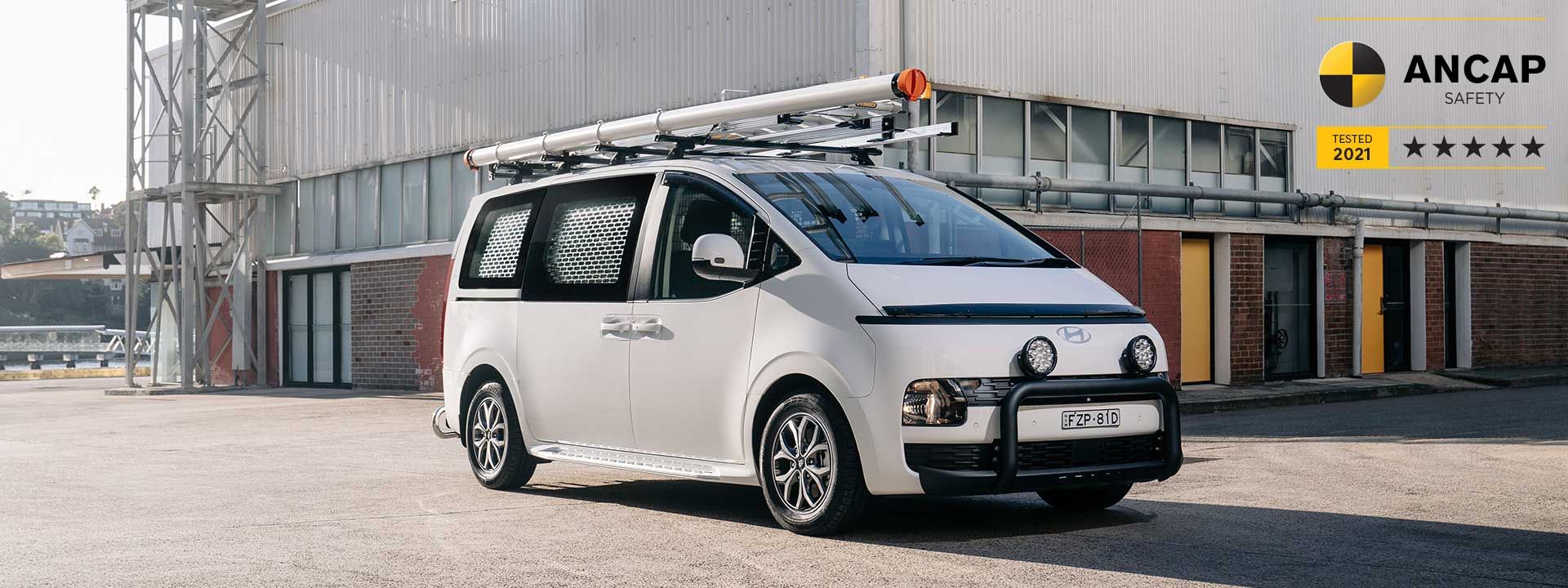 Hyundai STARIA Load commercial van parked outside an industrial building, showcasing practical cargo capabilities, modern design and a 5-star ANCAP safety rating.