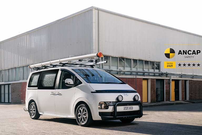 Hyundai STARIA Load commercial van parked outside an industrial building, showcasing practical cargo capabilities, modern design and a 5-star ANCAP safety rating.