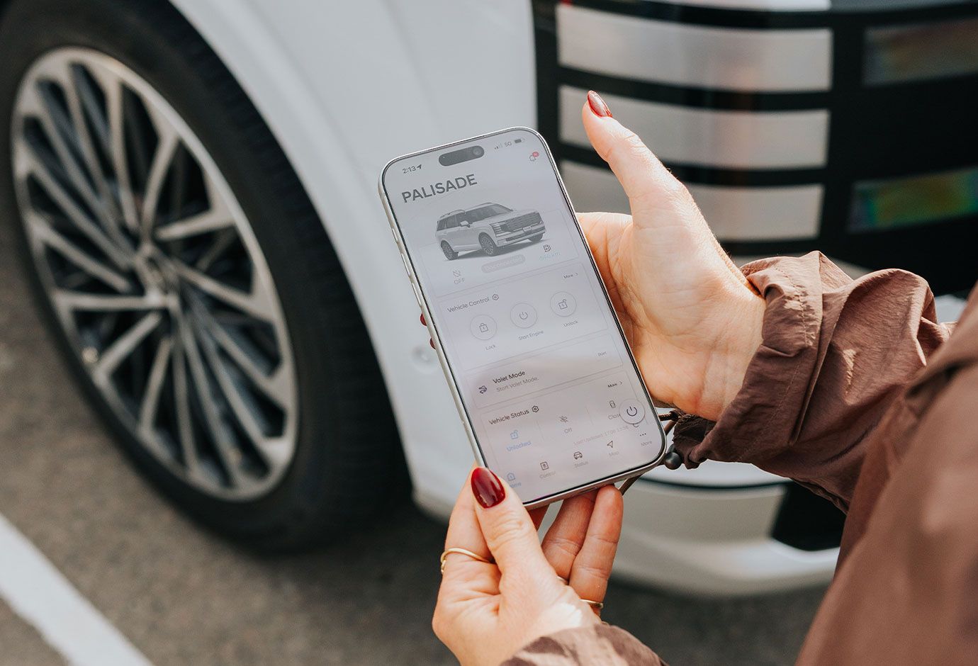 Person holding a smartphone displaying the Hyundai PALISADE Bluelink app, with the vehicle parked in the background.