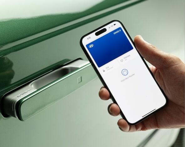 A person uses a smartphone with the Hyundai Digital Key app to unlock a green Hyundai IONIQ 9 by holding the phone near the vehicle's door handle.