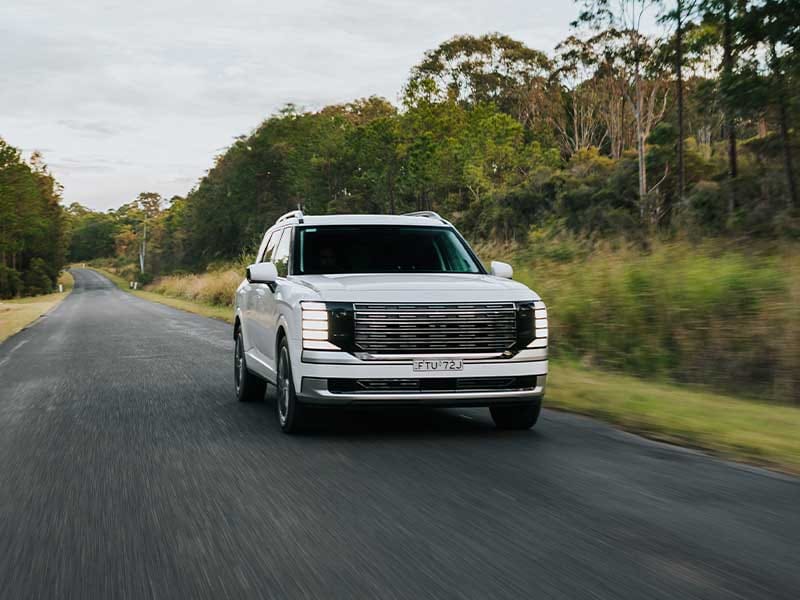 Hyundai PALISADE SUV driving along a scenic country road, showcasing its bold front grille, premium design, and commanding presence in a natural outdoor setting.