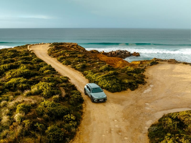 Hyundai Kona Hybrid driving along a scenic coastal dirt road with ocean waves in the background.
