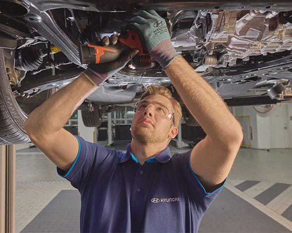 Hyundai technician performing a vehicle inspection and maintenance service underneath a Hyundai vehicle in a modern workshop, ensuring expert care, safety, and optimal performance.