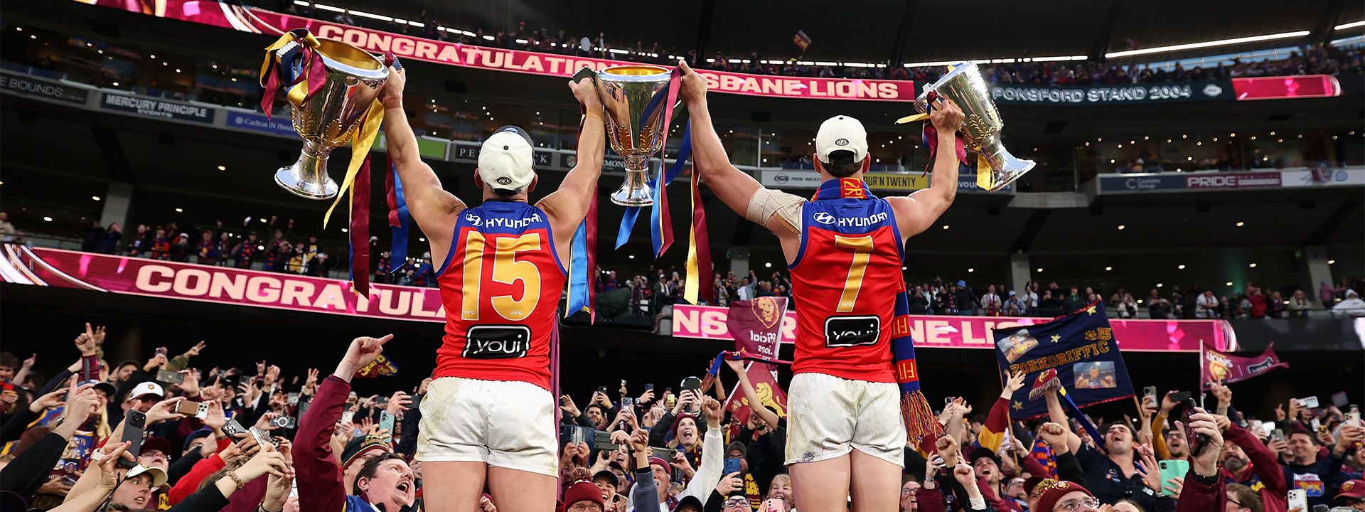 Brisbane Lions AFL Premiership celebration with players raising championship trophies in front of cheering supporters at a packed stadium, showcasing Hyundai’s partnership with the club.