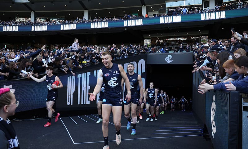 Hyundai-sponsored Carlton FC players run onto the field, engaging with fans at a packed AFL stadium, showcasing the partnership and matchday excitement.