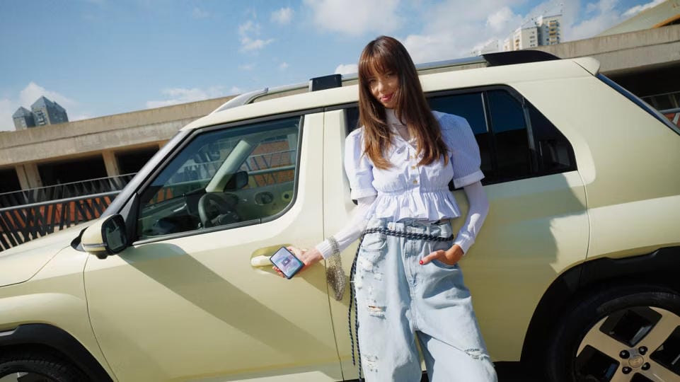 Una mujer se apoya en el nuevo coche eléctrico compacto Hyundai INSTER, utilizando Bluelink en su smartphone.