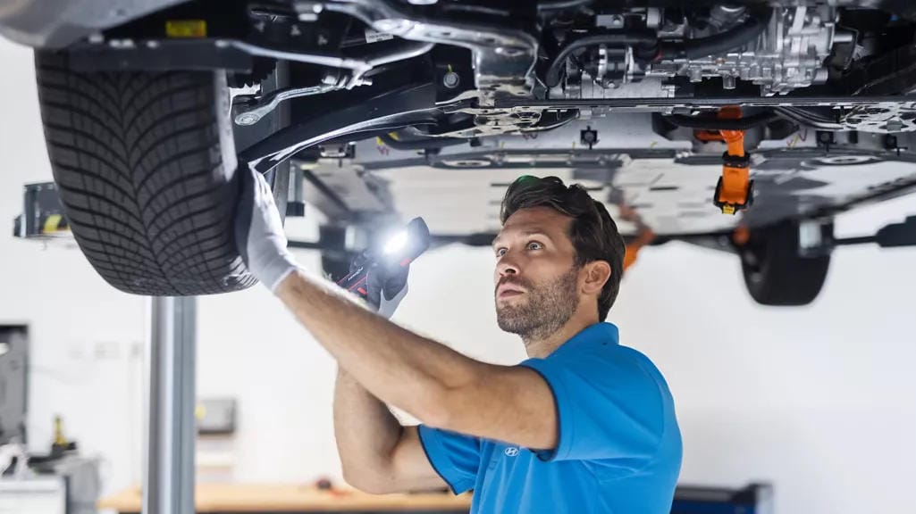 Fondo de estación de servicio vehicular. Mecánico realizando reparaciones y mantenimiento en un vehículo gris levantado.