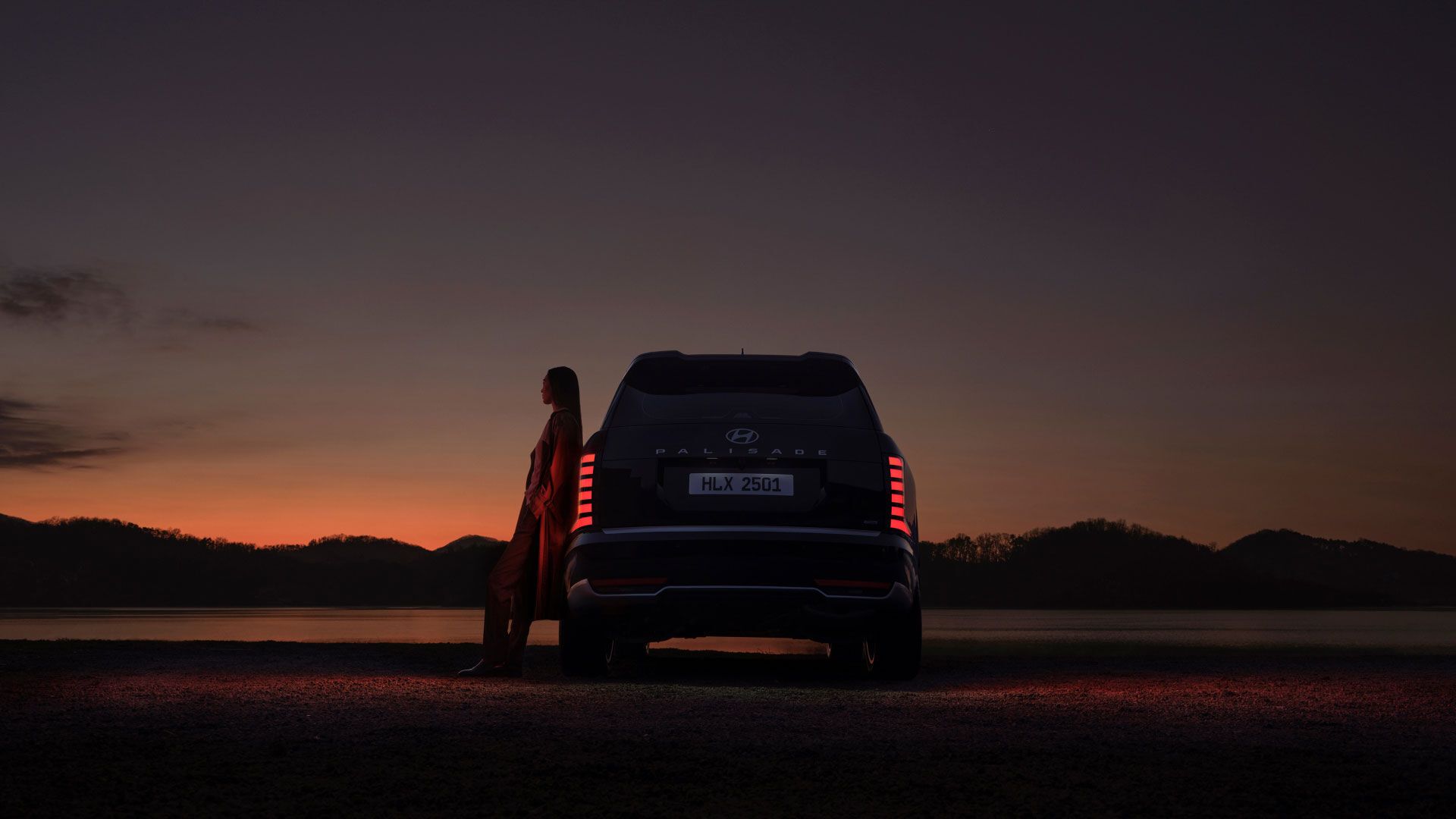 A woman leaning on the side of the all-new PALISADE, gazing at the sunset