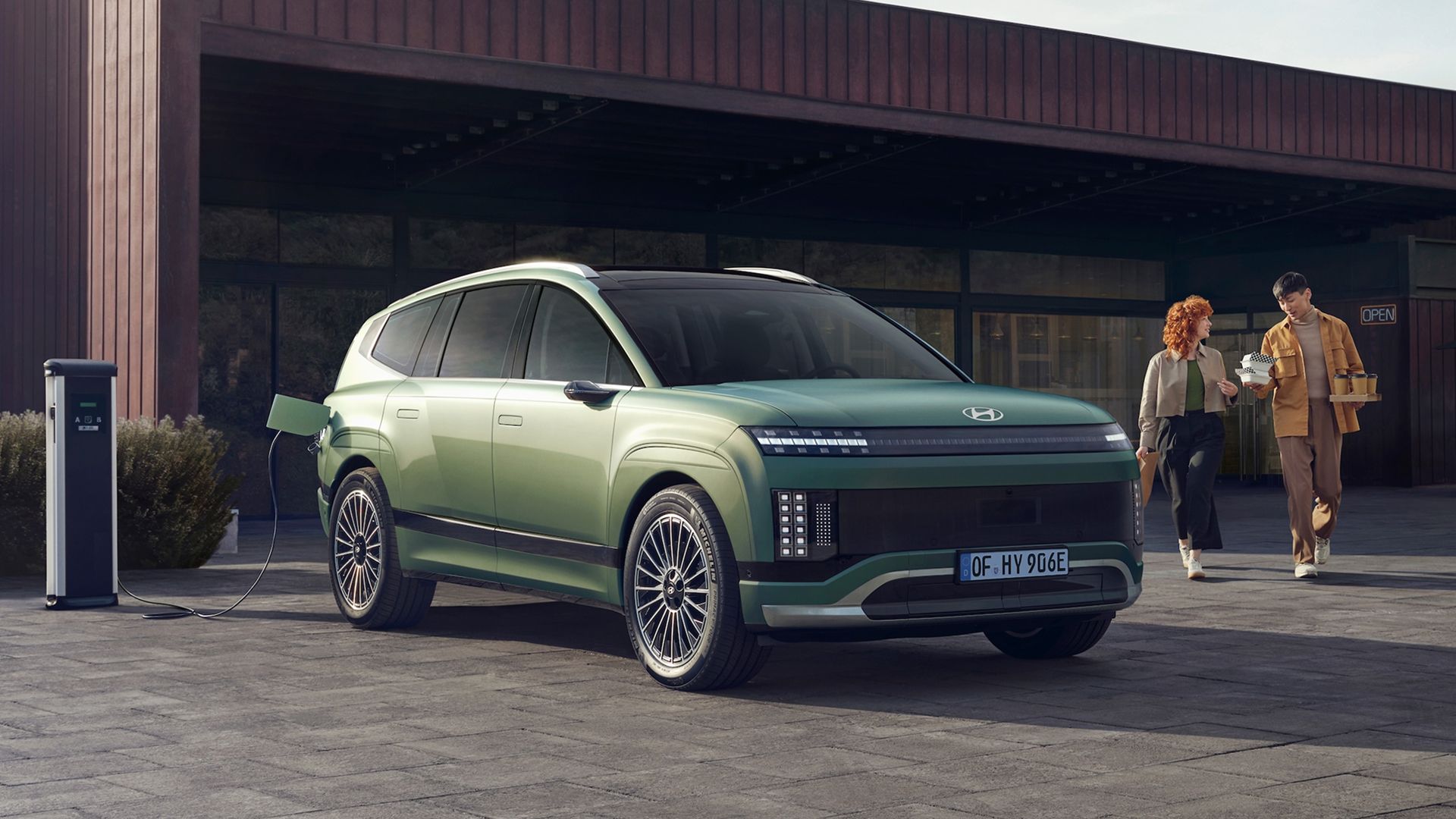Et ungt par kommer gående med kaffe bak en IONIQ 9 som står og lader. Foto.