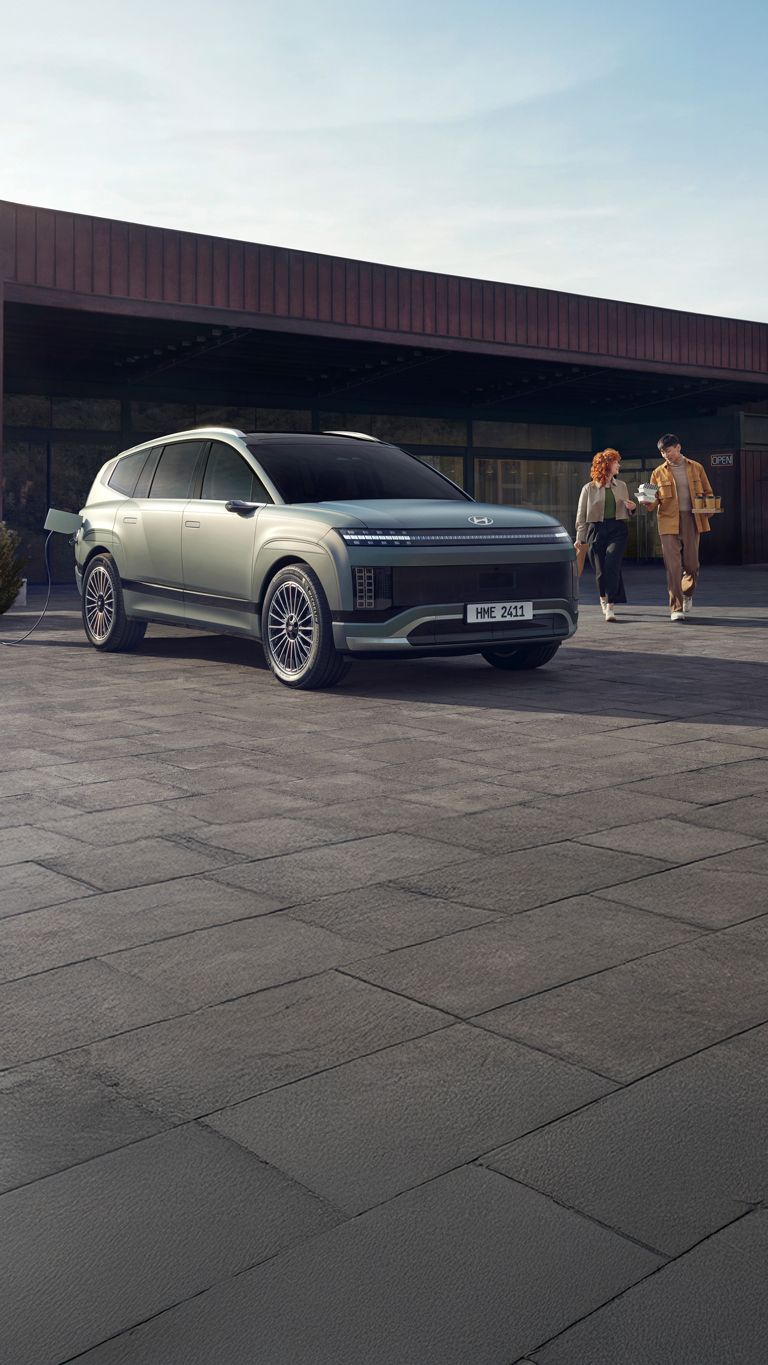 Et ungt par kommer gående med kaffe bak en IONIQ 9 som står og lader. Foto.