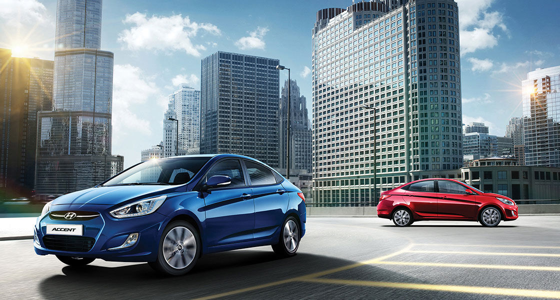 Side-front view of blue Accent in front and side view of red Accent parked at the parking lot with city view behind on a clear day