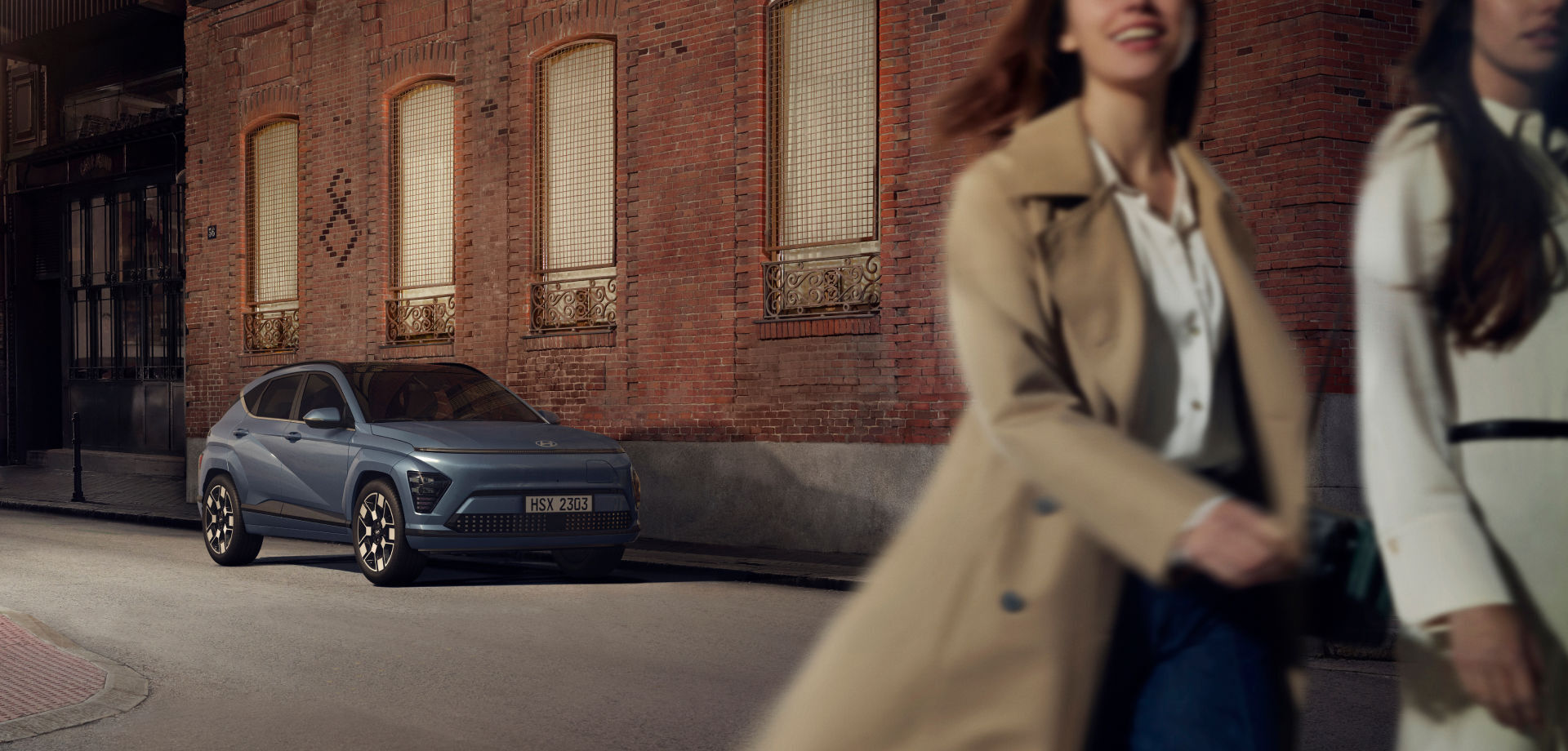 Two smiling women are walking behind the left front view of a parked The all-new KONA Electric in a dimly lit alleyway.