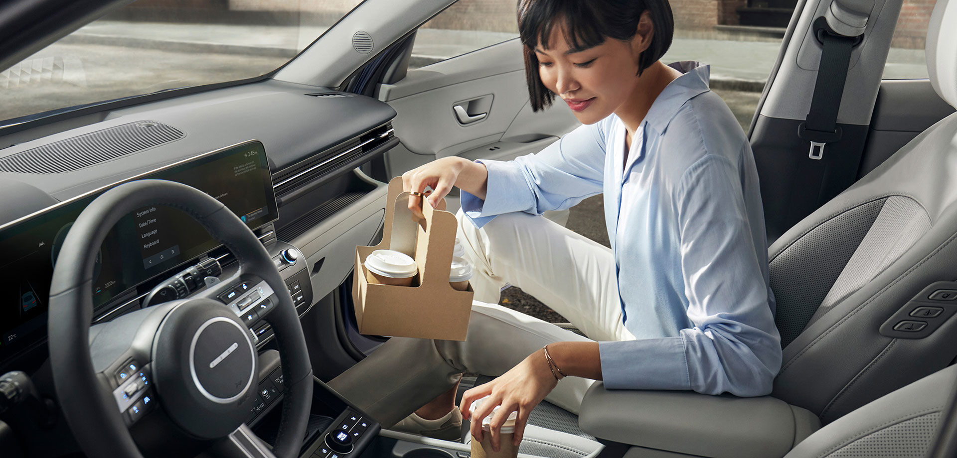 A woman is sitting in the passenger seat of The all-new KONA with the right front door open. The woman is holding a to-go drink in her right hand, and carrying her drink into a cup holder in her vehicle with her left hand.