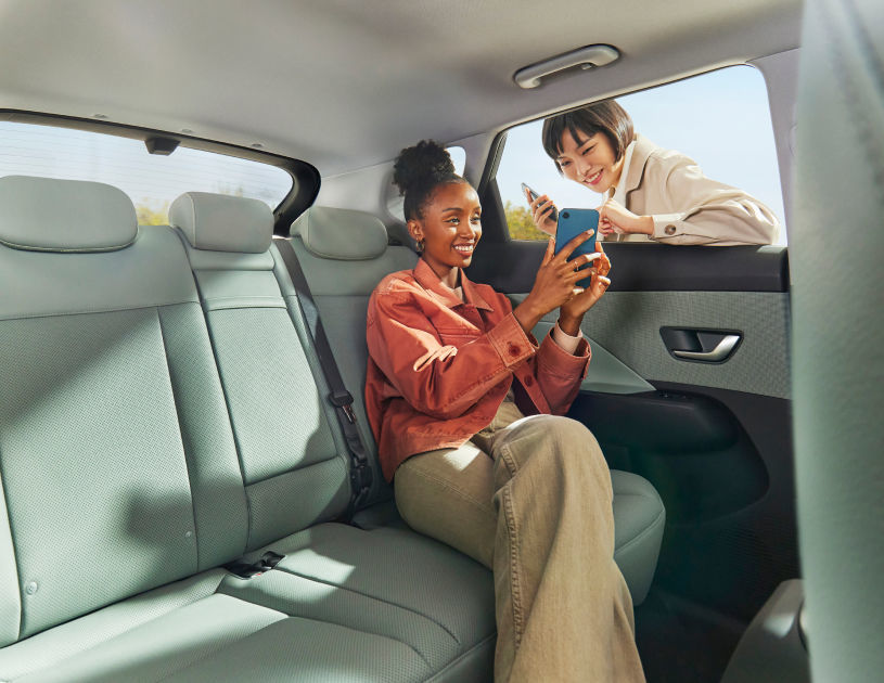 تجلس امرأة في الجزء الخلفي من السيارة كونا الجديدة كليًا تنظر إلى شاشة هاتف ذكي، بينما تظهر امرأة خارج السيارة من خلال نافذة مفتوحة.