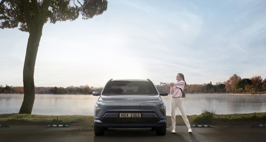 The front view of a The all-new KONA Electric standing in a parking lot by the lake. On the left side, a woman in sportswear is doing warm-up exercises.