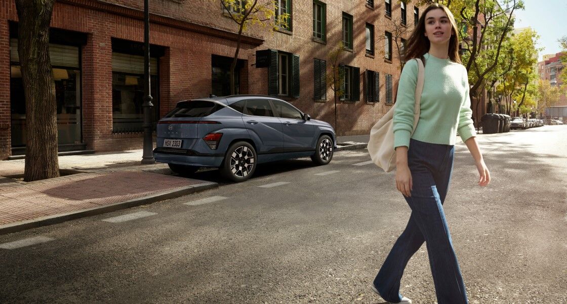 The rear view of a green The all-new KONA parked in a parking space on the roadside is focused in the center of the screen, and a woman crosses the road to the right of the car looking around.