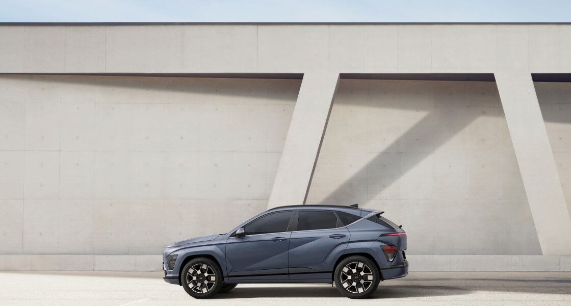 A blue The all-new KONA stands in front of a white wall with its left side facing up.