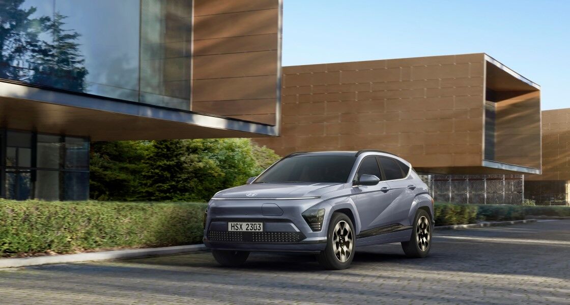 Left quarter view of a blue The all-new KONA Electric standing in front of a building with a distinctive rectangular exterior.