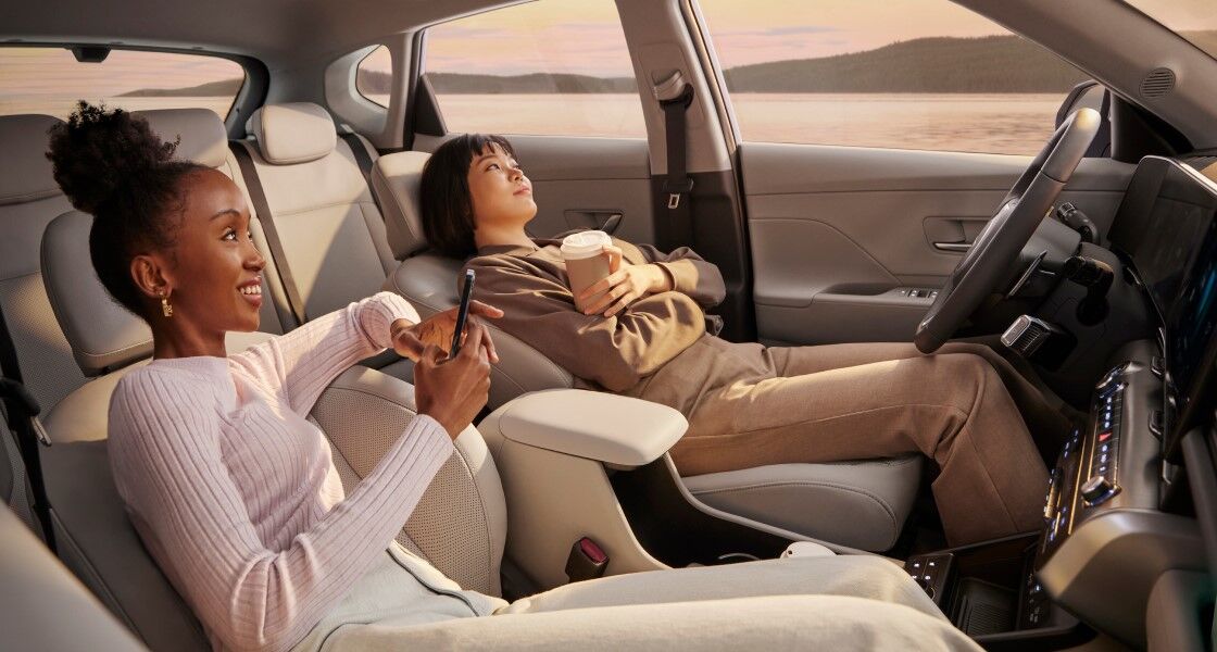 Two women are sitting in the front row of a The all-new KONA with their seats folded back, watching the sunset. The woman in the front passenger seat is holding her smartphone, and the woman in the driver's seat is holding her take-out paper cup in her left hand, with her arms crossed.
